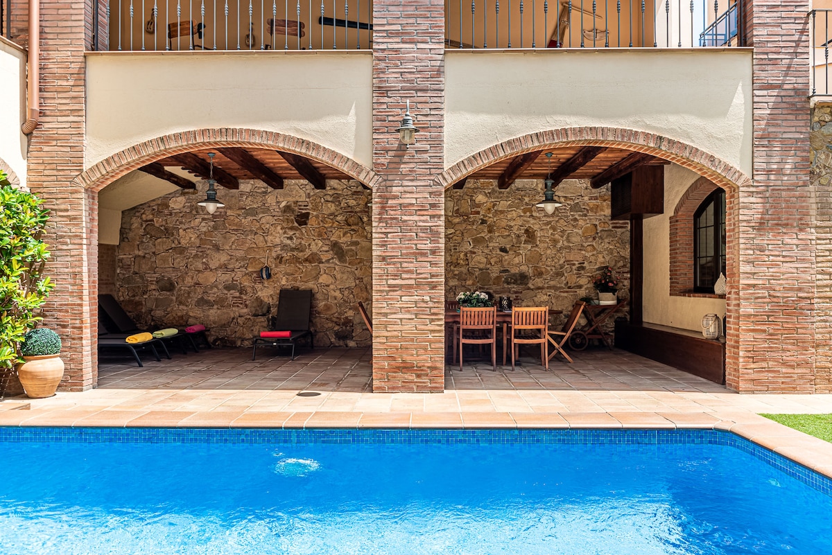 The outdoor area features a refreshing blue pool surrounded by a tile deck. Arched openings beneath a covered terrace reveal a rustic stone wall and wooden beams. Lounge chairs and a dining set are present, providing spaces for relaxation and meals outdoors.