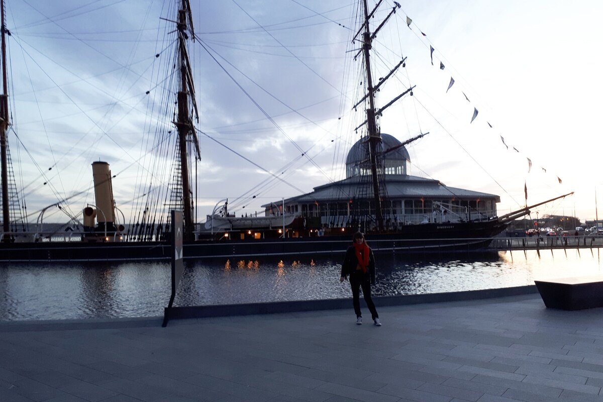 The image captures a historic ship docked near the waterfront, with its tall masts reaching upwards. The scene features a calm body of water reflecting the evening sky, and the nearby waterfront area is paved with smooth stones. The setting sun bathes the scene in soft light.