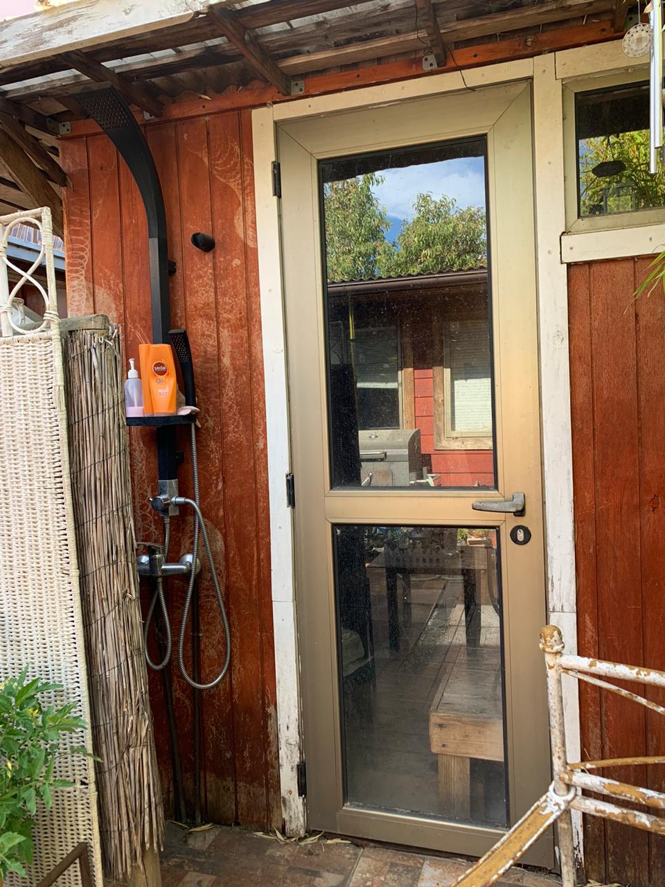 An outdoor shower is positioned next to a weathered wooden structure. A glass door allows views into a cozy indoor space, while a simple shower fixture is mounted on the exterior wall. Natural light filters through nearby greenery.