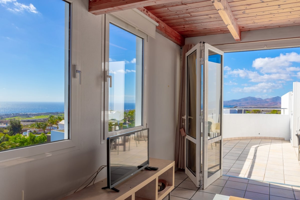 A bright living area is depicted with large windows showcasing unobstructed sea views. Natural light fills the space, complemented by wooden ceiling beams. A television is placed on a light wooden table, and open glass doors lead to a private terrace with outdoor seating.