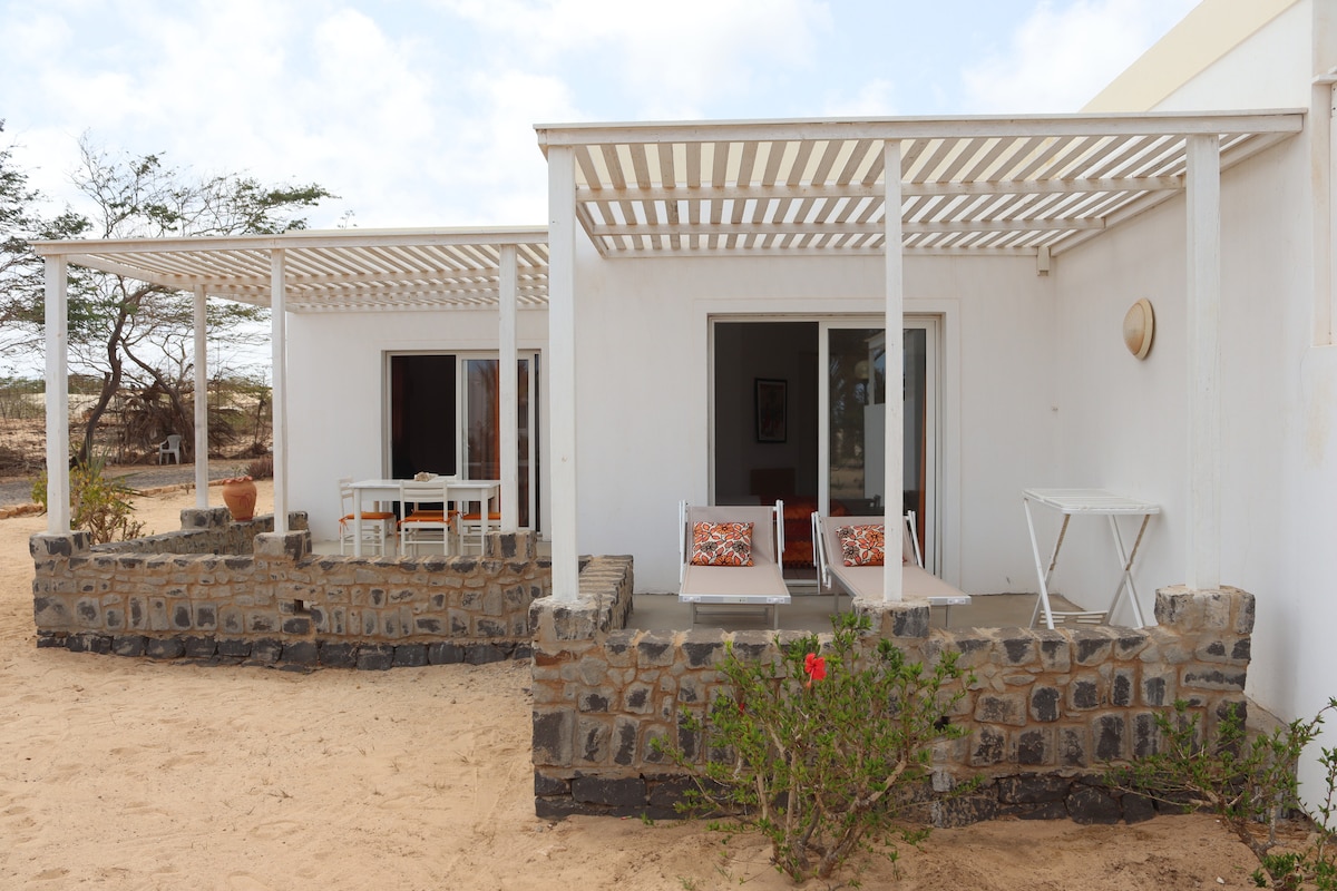 The exterior shows a house with white walls and a sandy area. A covered terrace with stripes provides shade over a seating arrangement, while chairs and a small table are visible. Bordering plants add a touch of greenery to the landscape.