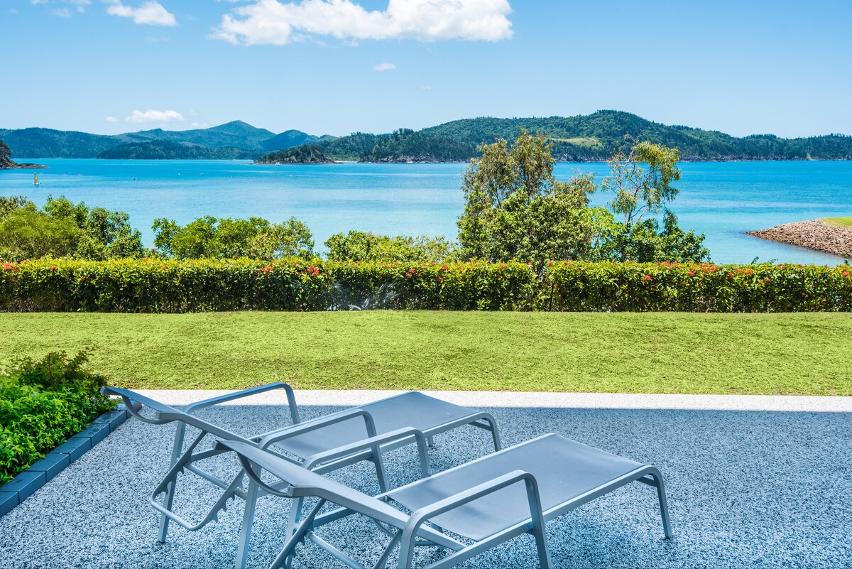 A clear view of the blue water and distant islands is framed by lush greenery. Two loungers are positioned on a patio, inviting relaxation while taking in the scenic landscape. The vibrant sky and gentle clouds enhance the tranquil outdoor setting.