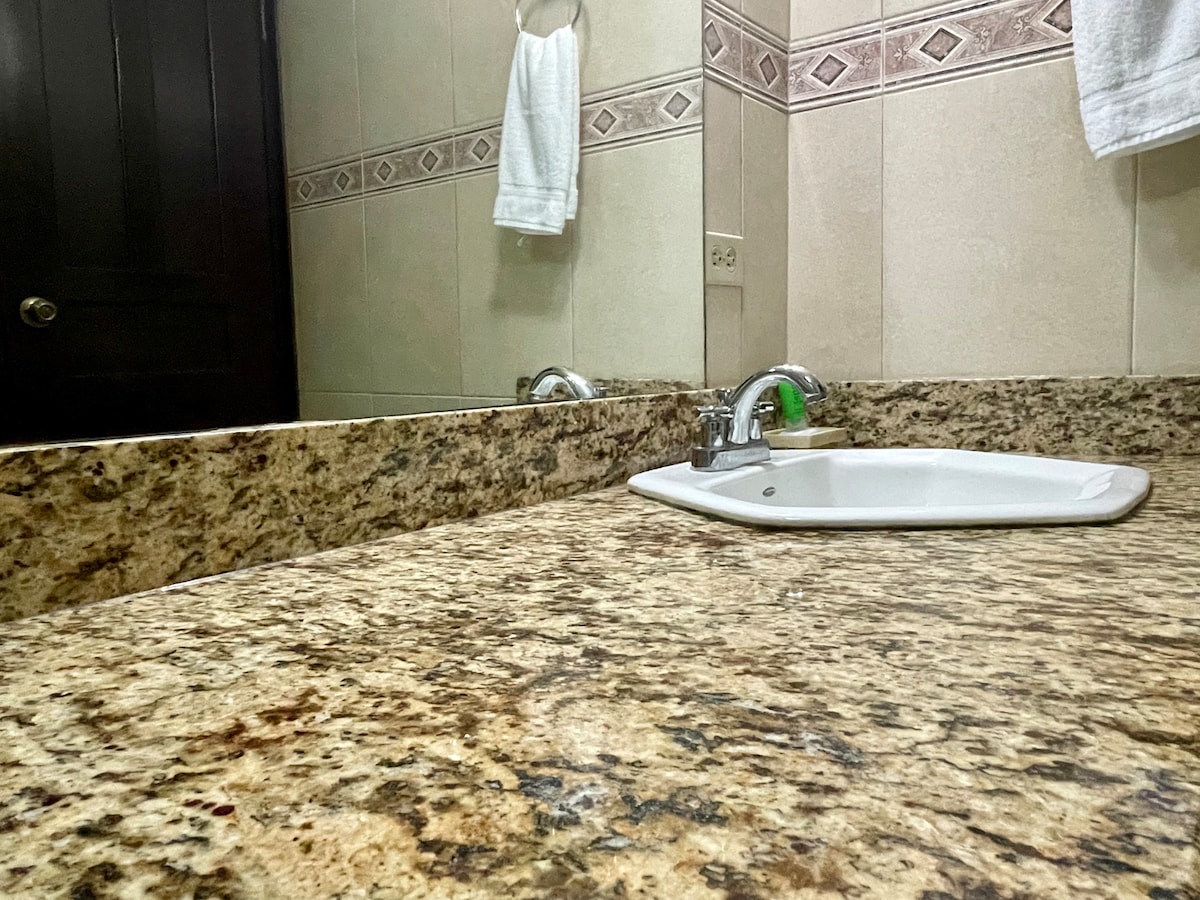 A clean and organized private bathroom features a granite countertop, a white sink, and a mirror reflecting the space. Fresh towels are neatly hung nearby, and the warm lighting enhances the overall ambiance.