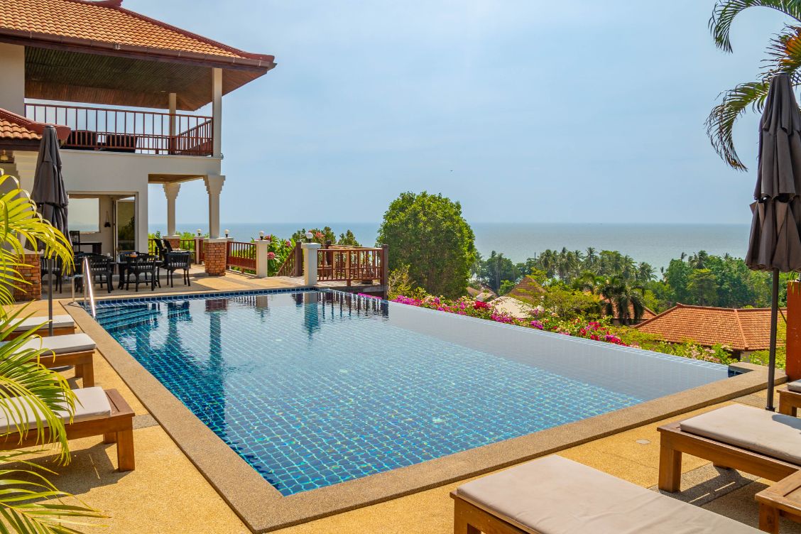 An infinity pool extends toward the horizon, offering views of the Andaman Sea. Comfortable loungers face the water, with umbrellas providing shade. The villa's upper level showcases a spacious outdoor seating area, surrounded by lush greenery and vibrant flowers.