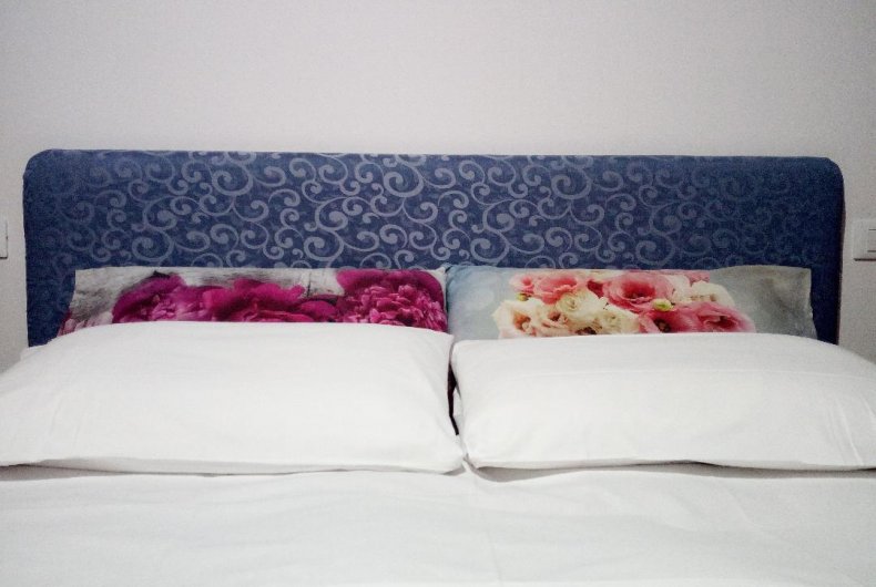 A spacious bed is presented with two large, plush white pillows. The headboard features a deep blue design, while floral accent pillows in shades of pink and cream add a touch of color.