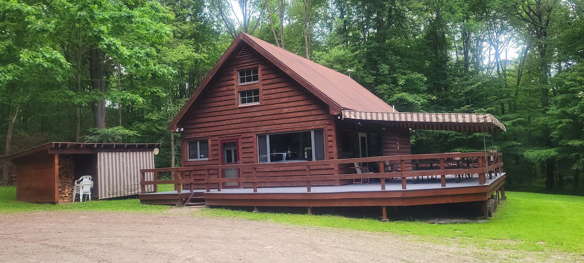A wooden cabin is surrounded by lush greenery, featuring a large attached deck with outdoor seating. A storage shed is visible nearby, and the well-maintained yard highlights the natural beauty of the wooded lot.