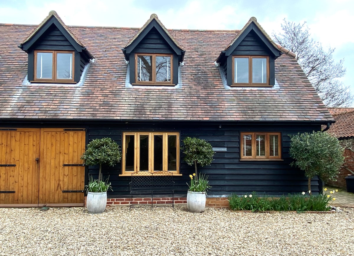 The exterior of Orchard End Barn is showcased, featuring a charming dark wooden façade with multiple windows. Two potted plants flank a central seating area adorned with a small table, surrounded by a gravel pathway. A wooden door offers access to the barn, enhancing its rustic appeal.