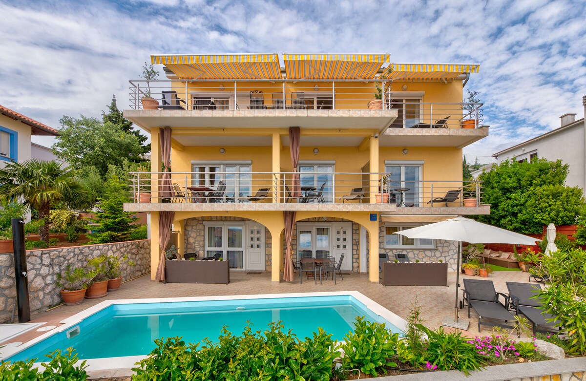 A three-story villa is showcased, featuring a pool area surrounded by lush greenery and potted plants. A canopy provides shade over the outdoor seating area. Spacious balconies with tables and chairs offer views of the surroundings, while large glass doors ensure plenty of natural light.