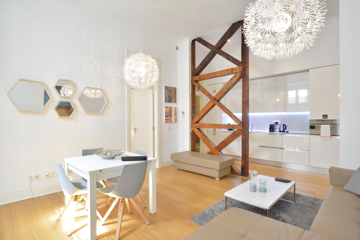 A stylish living and dining area features a light wooden floor, a white dining table set for six, and modern grey chairs. An inviting sofa with soft cushions sits nearby. Unique light fixtures add a touch of elegance, complemented by wooden beams enhancing the open layout.