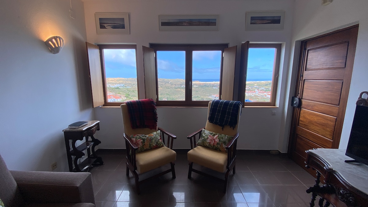 A cozy seating area is presented with two wooden armchairs facing large windows, offering expansive views of the valley. Soft throw blankets are draped over the chairs, which rest on polished tile flooring. A small side table sits beside the armchairs, complementing the natural light entering the space.