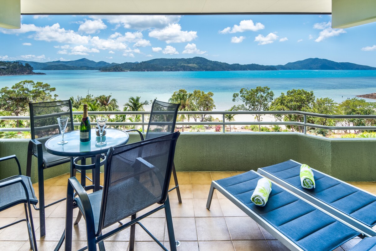 A balcony area features an outdoor dining table with four black chairs and a glass top. A sun lounge rests adjacent to the table, adorned with rolled towels. Breathtaking views of the beach and water are visible in the background, framed by lush greenery.