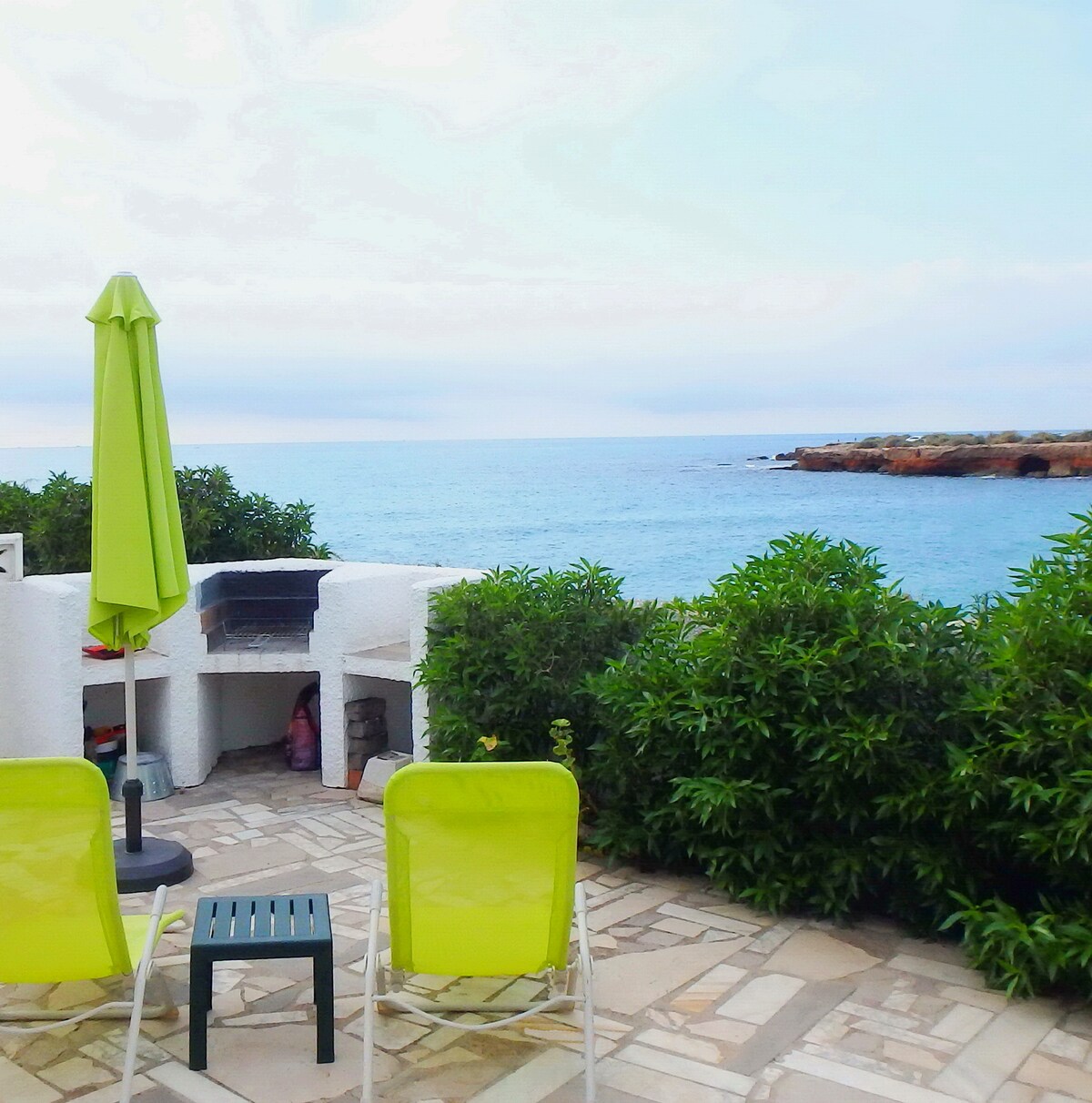 A terrace area overlooks the calm sea, featuring a barbecue area and vibrant green shrubs. Two loungers with umbrellas provide comfortable seating for relaxation, while the gentle waves of the ocean create a serene backdrop.