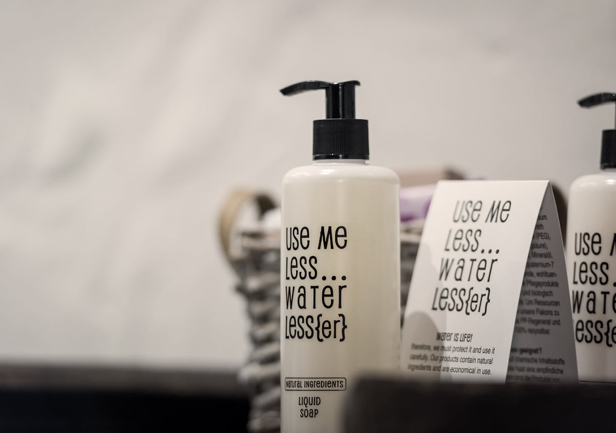 A liquid soap bottle is displayed prominently on a wooden surface. The label features clear, bold text with the instruction 'Use Me Less...Water Less(er).' Next to the bottle, a folded informational card provides details about its ingredients and usage. A woven basket holds the items, adding a natural texture.