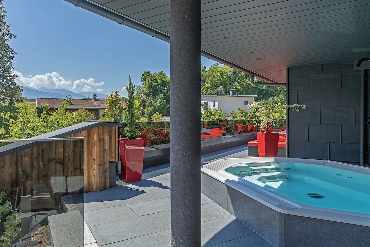 An outdoor terrace is featured, with a luxurious hot tub surrounded by elegant stone tiles. Bright red lounge chairs are positioned nearby, offering relaxation amidst lush greenery. The view extends to the landscape beyond, with partially cloudy skies enhancing the serene atmosphere.