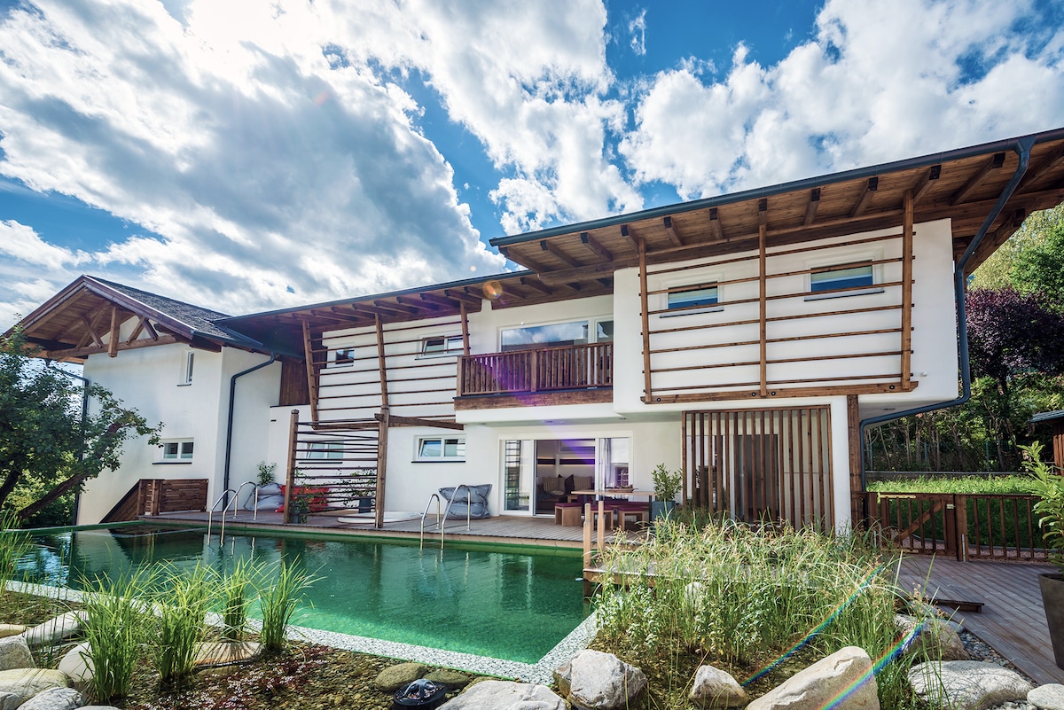 A modern, architecturally striking house is set beside a natural pool, surrounded by lush greenery. The structure features wooden accents and spacious balconies, with large windows reflecting the sky. The tranquil pool area is complemented by carefully arranged stones and aquatic plants.