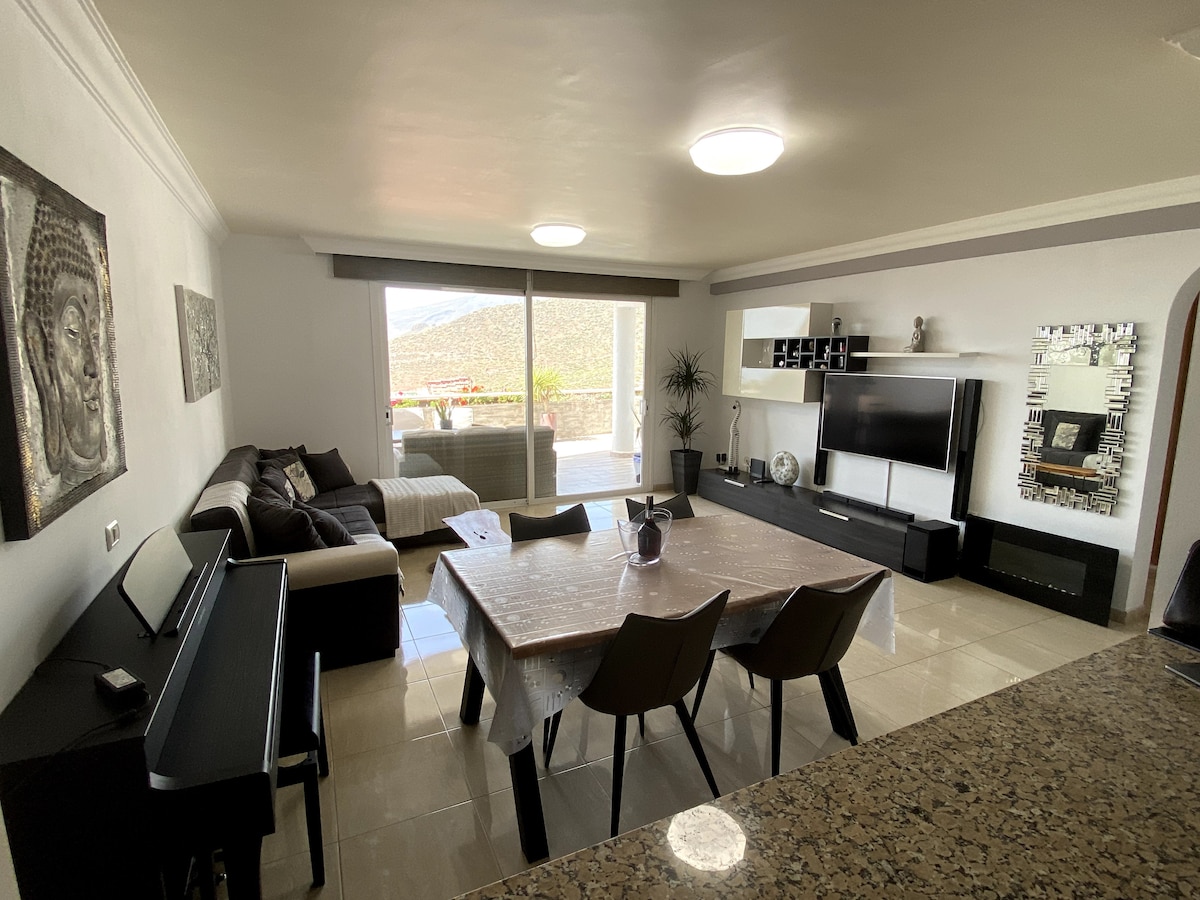 A modern living area features a spacious seating arrangement with a dark sofa and patterned cushion, complemented by a contrasting light rug. A dining table is set for six, while large windows provide natural light and a view of the terrace outside.