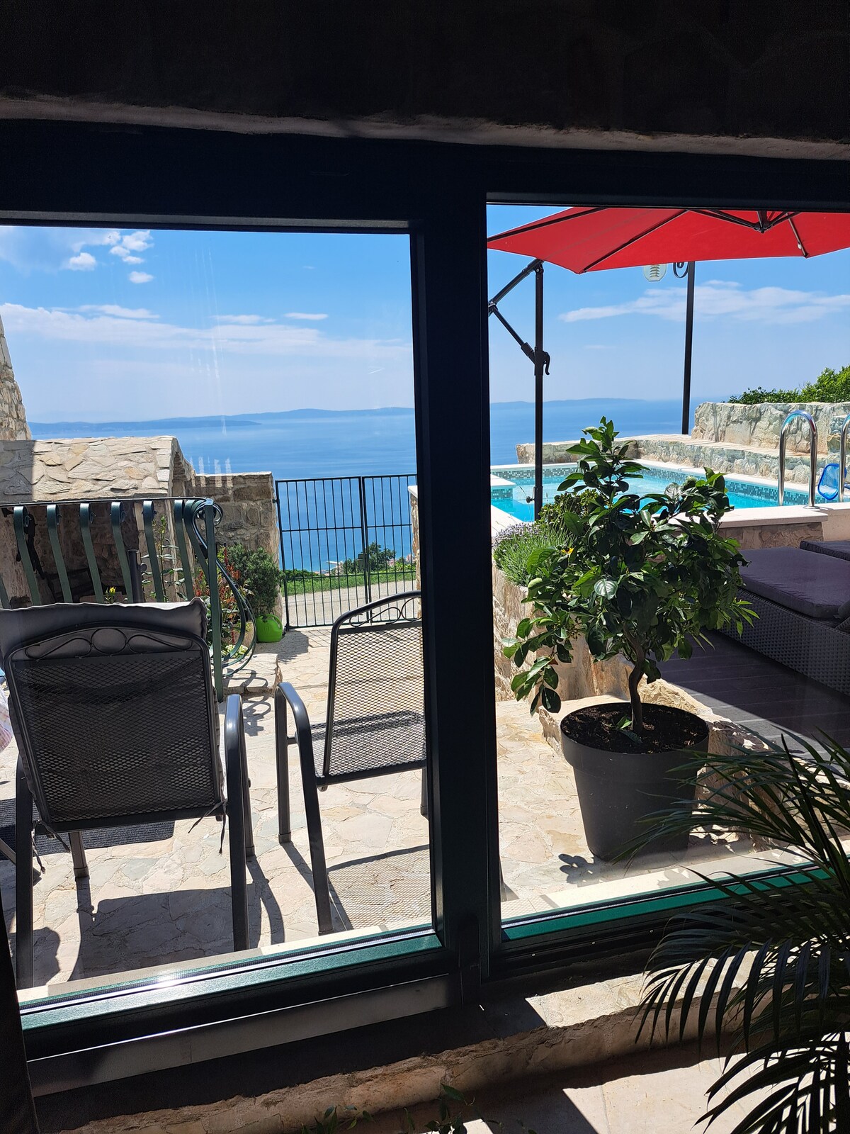 The image displays a panoramic view of the sea through large glass doors. Outdoor seating is visible, including a table and chairs under a shaded area. A lush green plant adds a touch of nature, framing the scene with the swimming pool and stunning landscape beyond.