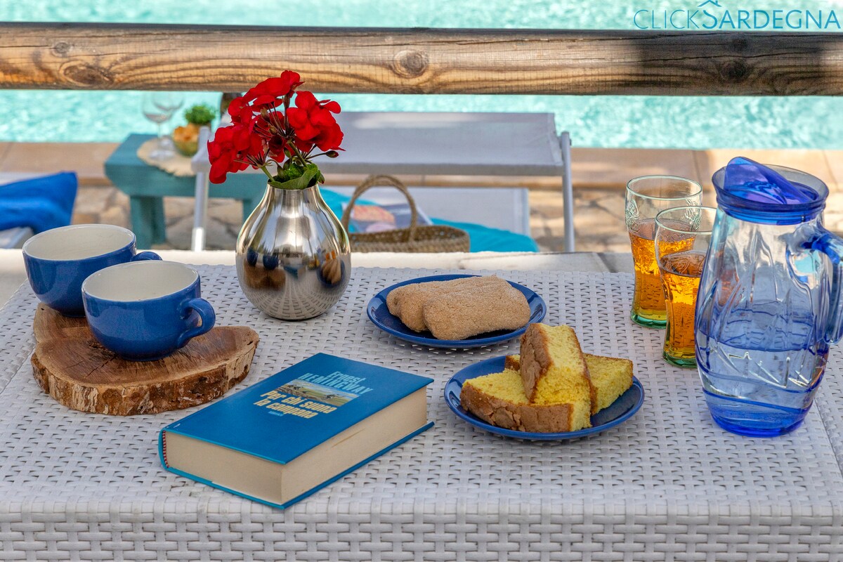 A table setting is displayed with a vibrant flower arrangement and two blue cups. Freshly baked treats are arranged alongside a book, a glass pitcher, and two glasses. The background features a sparkling pool, contributing to a relaxing ambiance.