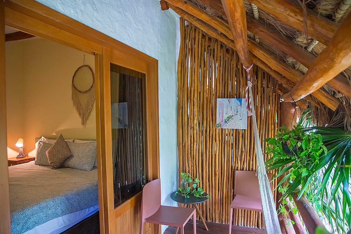 A cozy balcony is adorned with tropical plants and a small round table with two pink chairs. The entrance to a bedroom is visible, featuring a spacious bed and soft lighting, complemented by natural wood beams and woven accents on the walls.