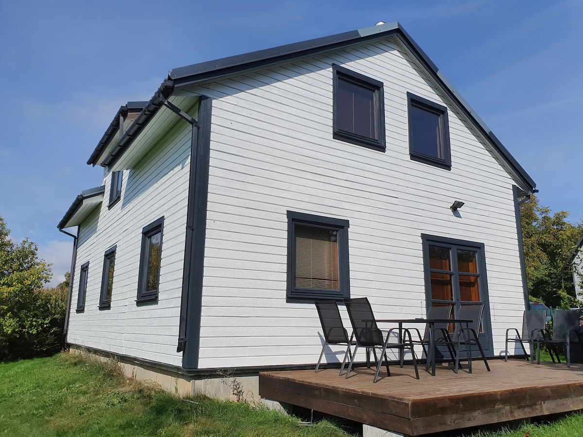 A two-story house with a white exterior and dark trim is surrounded by a green lawn. A wooden deck features outdoor seating arrangements. Large windows are present on both floors, allowing natural light to enter the interior.