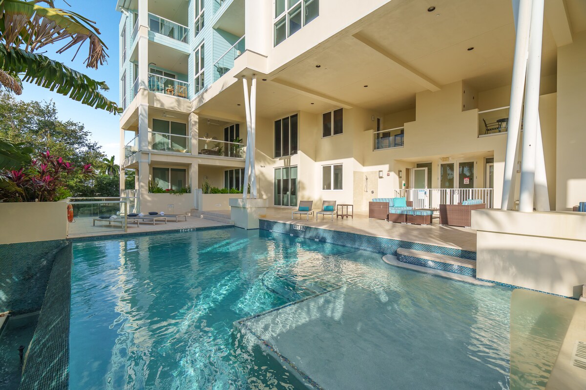 A modern infinity pool glistens under the sun, surrounded by elegant landscaping and sheltered patio areas. Comfortable seating options are positioned near the water, inviting relaxation. The building's contemporary architecture is showcased with large windows reflecting the clear blue sky.