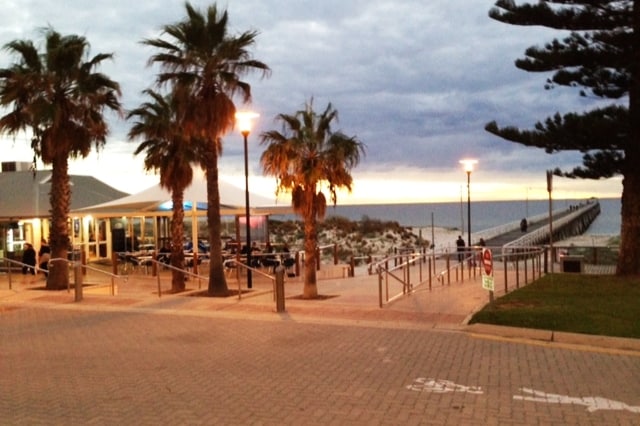 A beachfront scene captures palm trees lining a walkway leading to a restaurant with outdoor seating. The pathway extends towards a jetty, while the horizon displays gentle waves under a cloudy sky, hinting at sunset with warm tones in the background.
