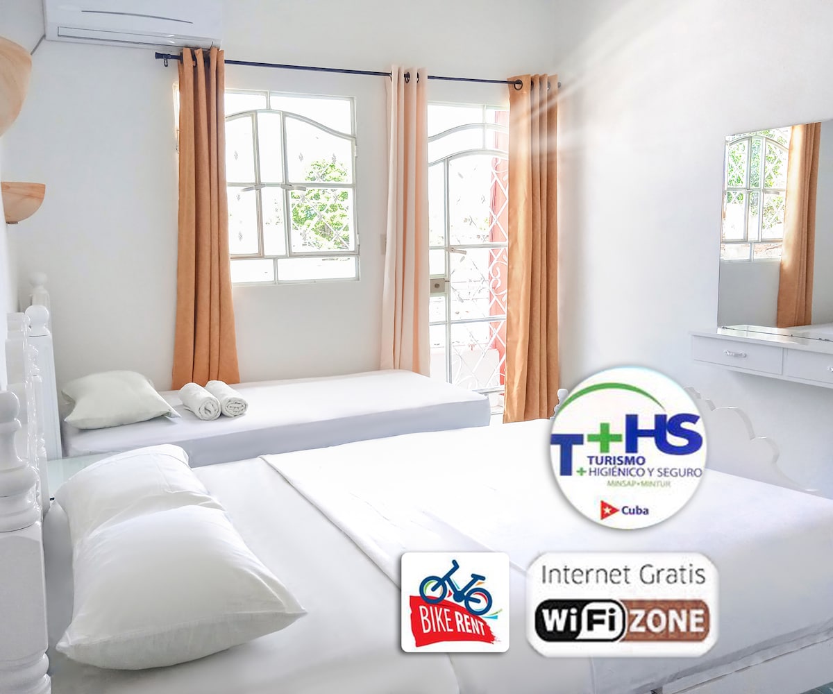 A bright room is presented with two beds, one spacious and the other a smaller size. Large windows allow natural light to fill the space, complemented by light-colored curtains. A mirror reflects the room’s simplicity and cleanliness, while neatly arranged towels are placed on the beds.