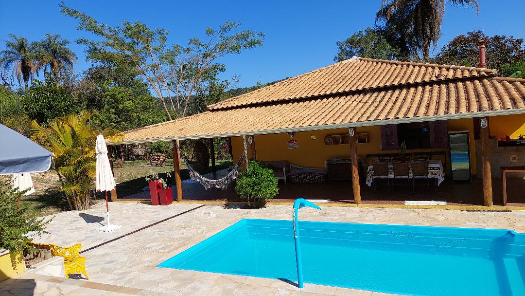 A sunlit outdoor area features a sparkling pool surrounded by a tiled deck. An open-air kitchen and dining space can be seen under a spacious wooden roof. Lush greenery and palm trees create a natural backdrop, while a hammock is hung in a shaded corner for relaxation.