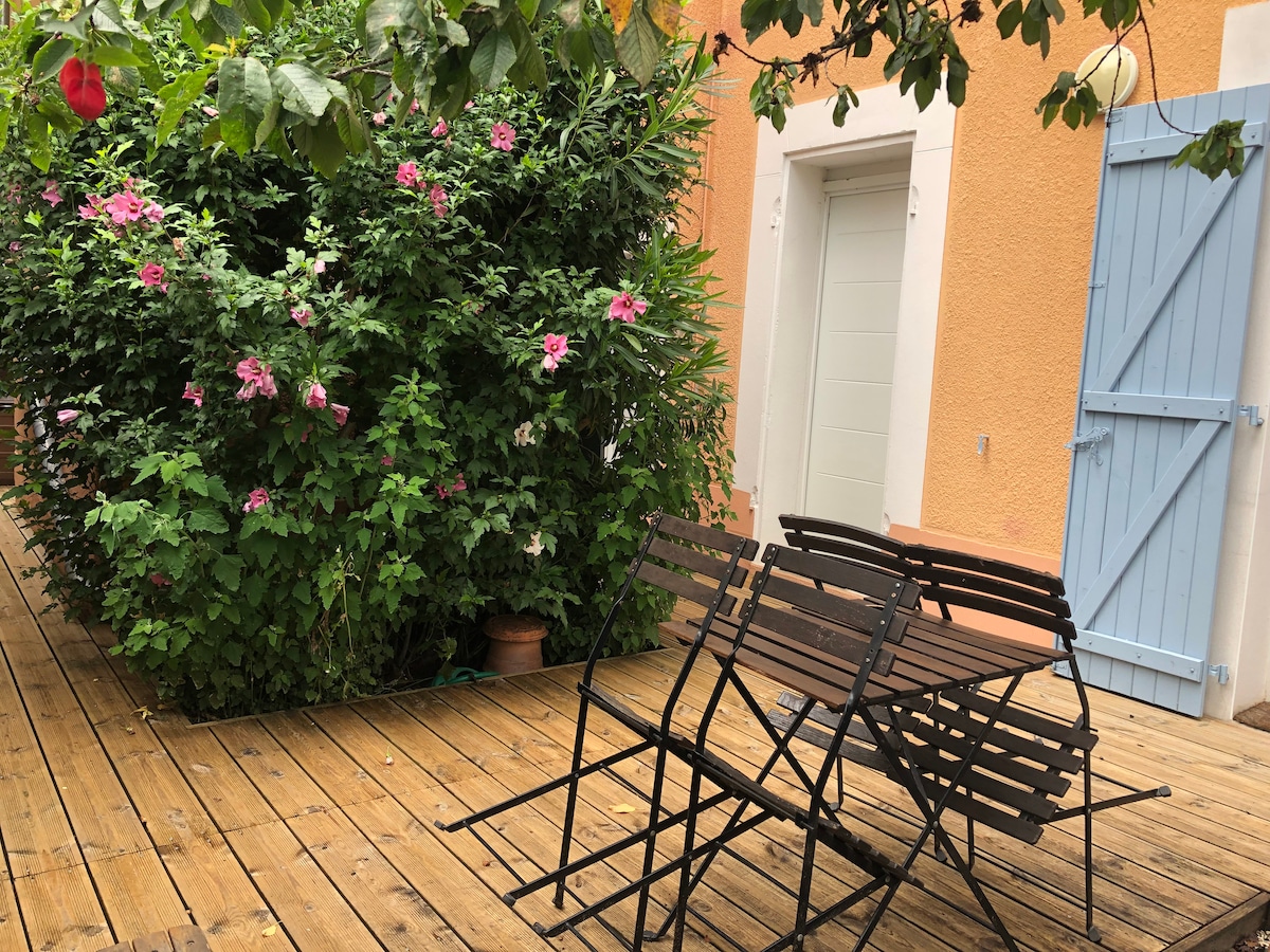 A wooden terrace is presented, featuring folding metal chairs arranged neatly. Lush greenery and vibrant pink flowers surround a doorway, enhancing the outdoor space's natural charm. The warm tones of the wall complement the serene atmosphere, suggesting a peaceful area for relaxation.