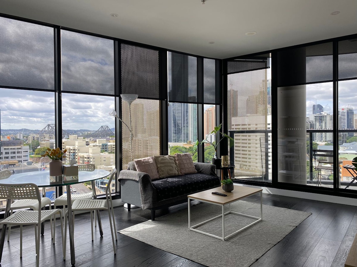 A stylish lounge area is showcased with large floor-to-ceiling windows displaying captivating views of the Brisbane skyline and Story Bridge. A comfortable sofa, light wooden coffee table, and dining chairs create an inviting space, complemented by soft natural light filtering through sheer blinds.