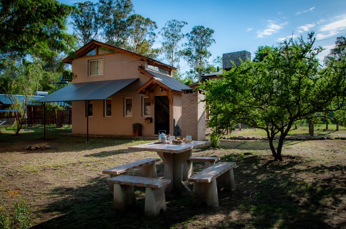 A cozy cabin with a sloped roof is surrounded by lush greenery. A wooden picnic table is positioned in front, complemented by stone seating. Sunlight filters through the trees, creating a peaceful outdoor environment for relaxation.