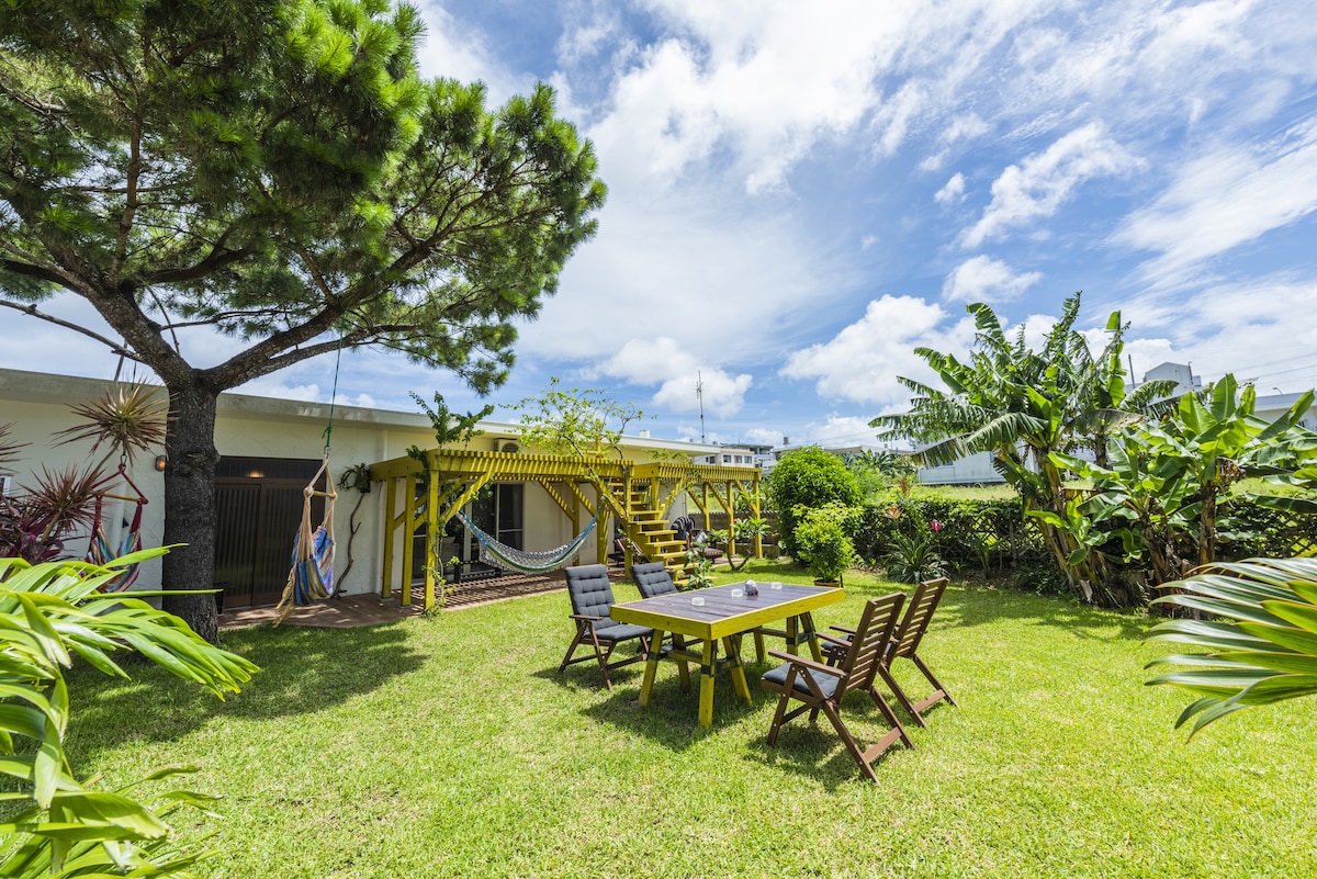 A spacious outdoor area features a large table surrounded by wooden chairs, with a hammock hanging nearby. Lush greenery, including tropical plants and trees, surrounds the yard, providing a serene environment for relaxation. The bright sky completes this inviting scene.