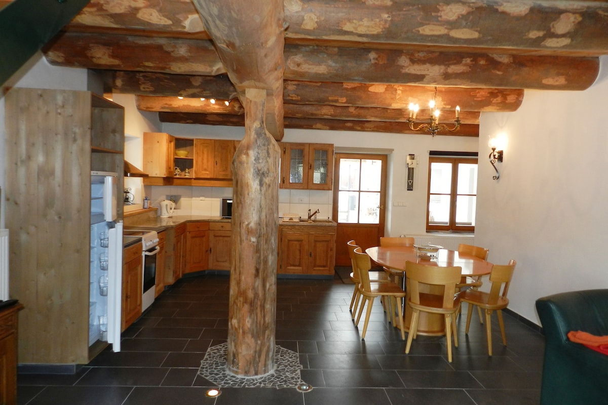 A spacious kitchen and dining area feature rustic wooden beams overhead and a central support column. The kitchen is equipped with modern appliances, while a round dining table surrounded by wooden chairs invites shared meals. Natural light enters through the windows, illuminating the dark flooring.