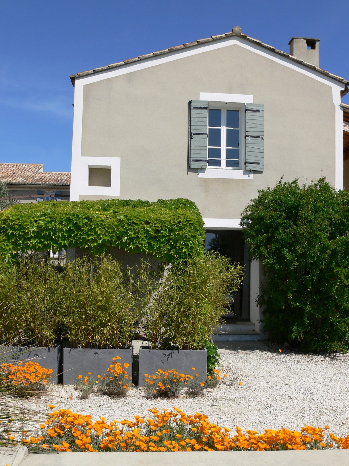 The exterior of a two-story house is shown, featuring a pale facade with green shutters. Lush greenery surrounds the entrance, complemented by vibrant orange flowers planted in rectangular planters, enhancing the inviting appearance of the property.