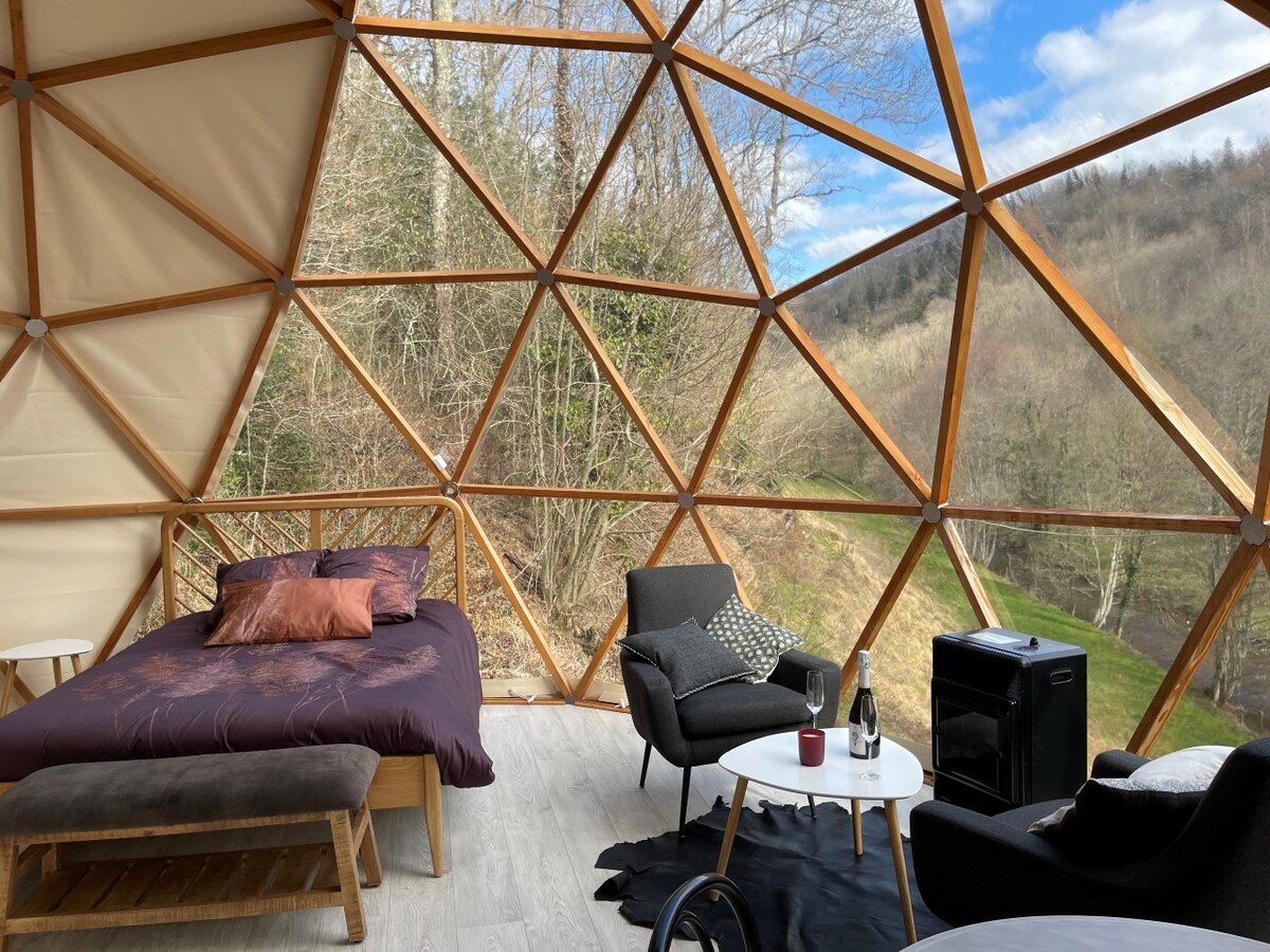 The interior of the geodesic dome features a spacious bed dressed in deep purple linens, accompanied by two cozy armchairs. A small table holds drinks, while a mini-fridge is positioned next to the seating area, all framed by panoramic windows that offer views of the surrounding nature.