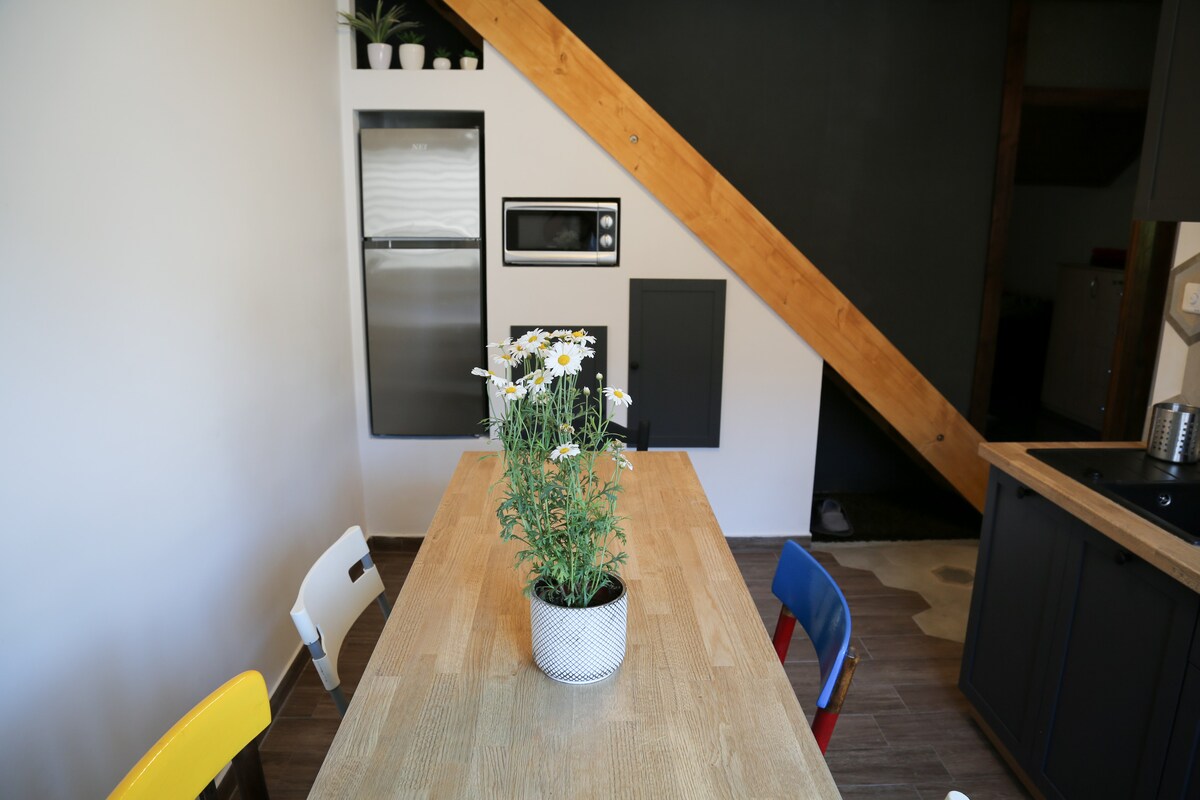A dining area features a wooden table surrounded by four chairs in varying colors. A vase with fresh daisies acts as a centerpiece. The space is complemented by modern appliances and a contrasting black wall, accentuated by the warm tones of the wooden beams.