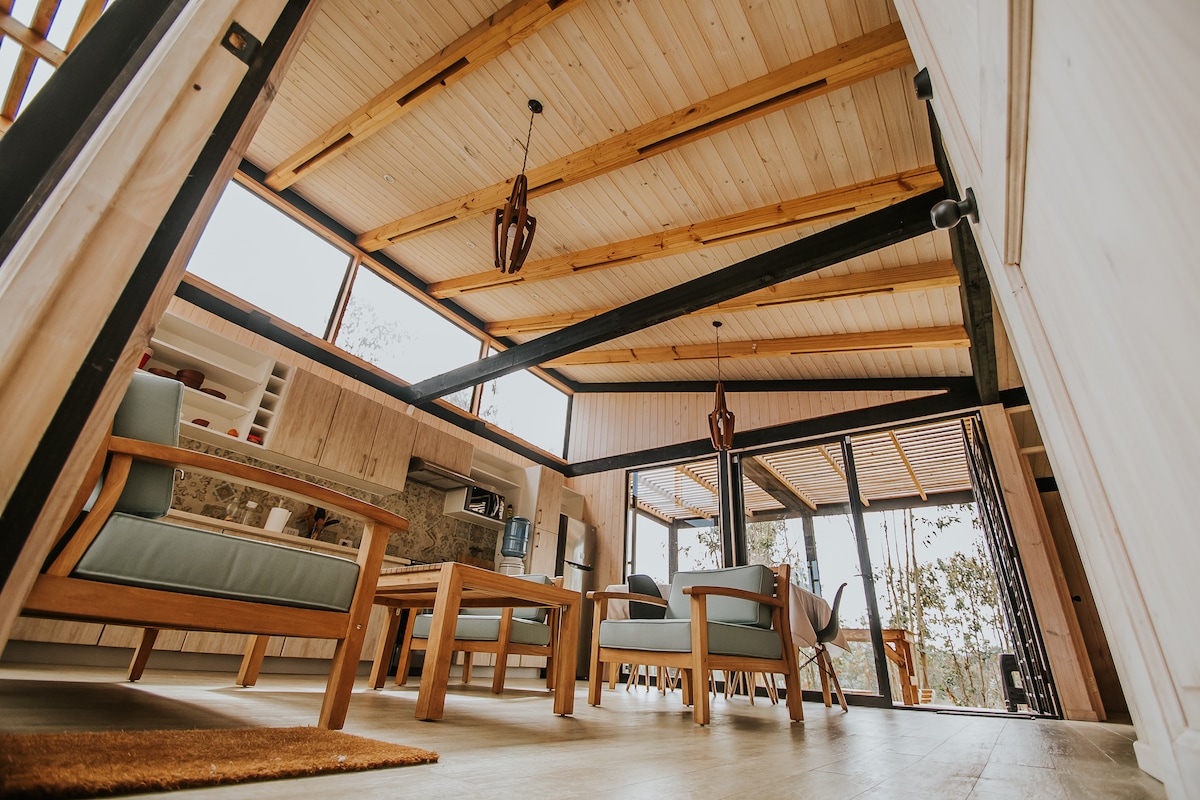The open living area features wooden beams and large windows that provide ample natural light. Comfortable seating arrangements, including two armchairs and a central table, create a relaxed space. A kitchenette is visible in the background, complementing the inviting design.