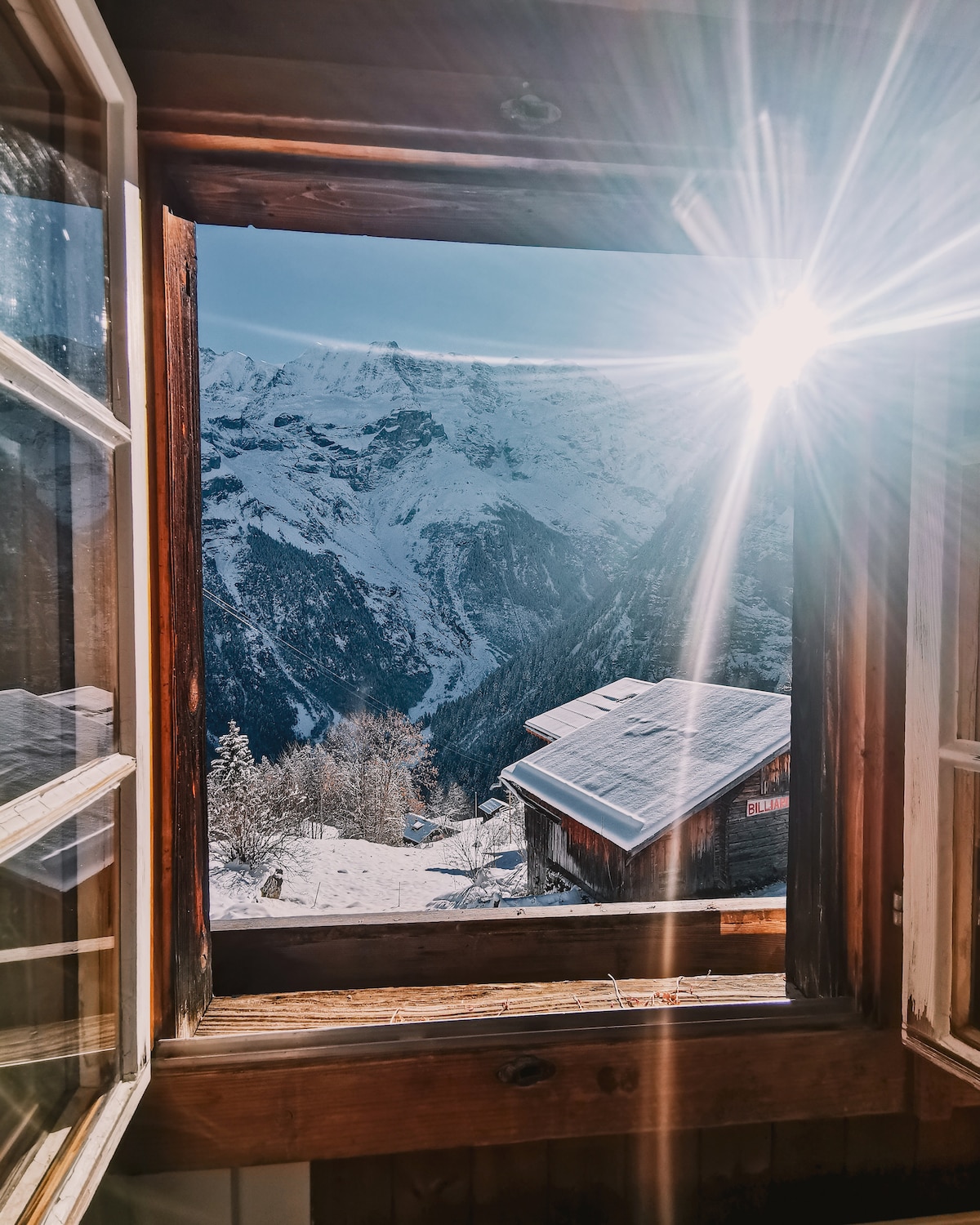 A window is shown with wooden frames, revealing a stunning view of snow-covered mountains and a distant cabin. Sunlight streams in, creating a bright and warm atmosphere inside, while the natural landscape showcases the serene beauty of winter in the mountains.