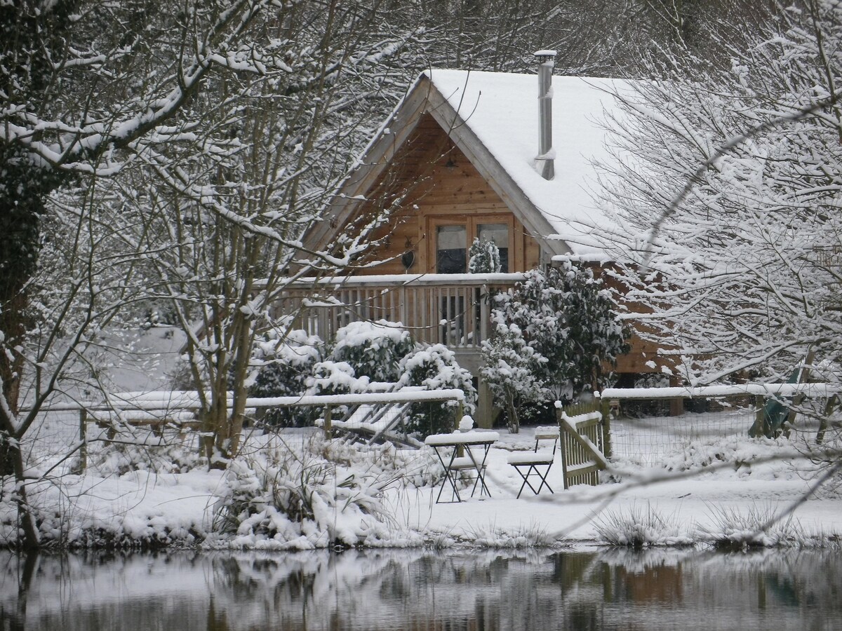 A wooden cabin nestled in a serene winter landscape, surrounded by trees blanketed in snow. A small terrace with two chairs overlooks a tranquil pond, reflecting the snowy scene. A chimney emerges from the cabin, suggesting warmth inside.