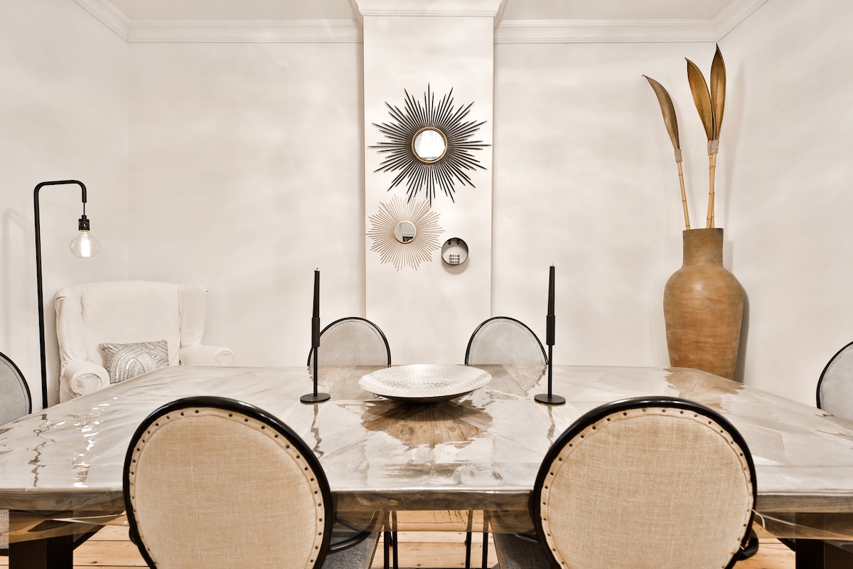 The dining area features a large table surrounded by rounded chairs with light fabric. Two slender candles stand on the table, and a decorative plate is centered. A striking mirror arrangement and a tall vase with decorative elements are displayed on the walls, enhancing the room's character.