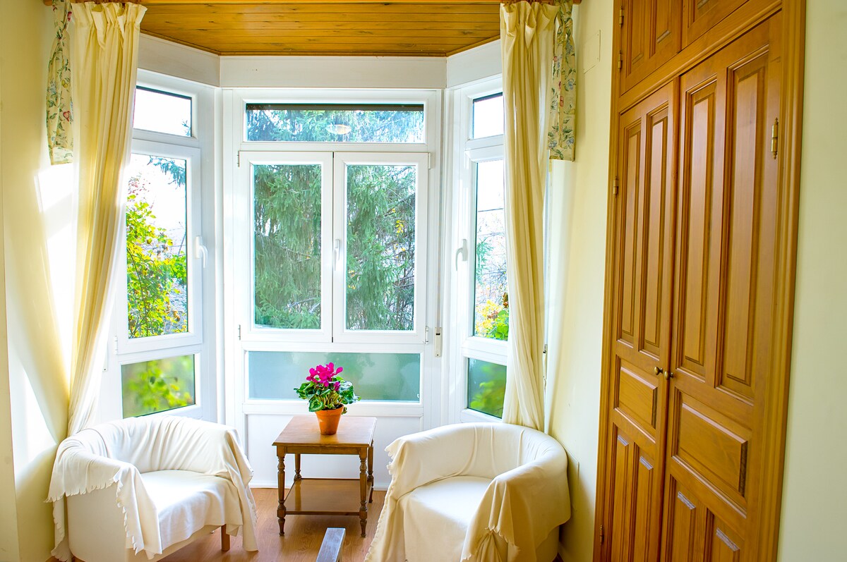 A bright and airy seating area features two upholstered chairs flanking a small wooden table. Tall windows allow natural light to fill the space, with sheer curtains framing the views of the greenery outside. A potted plant adds a touch of color to the room.