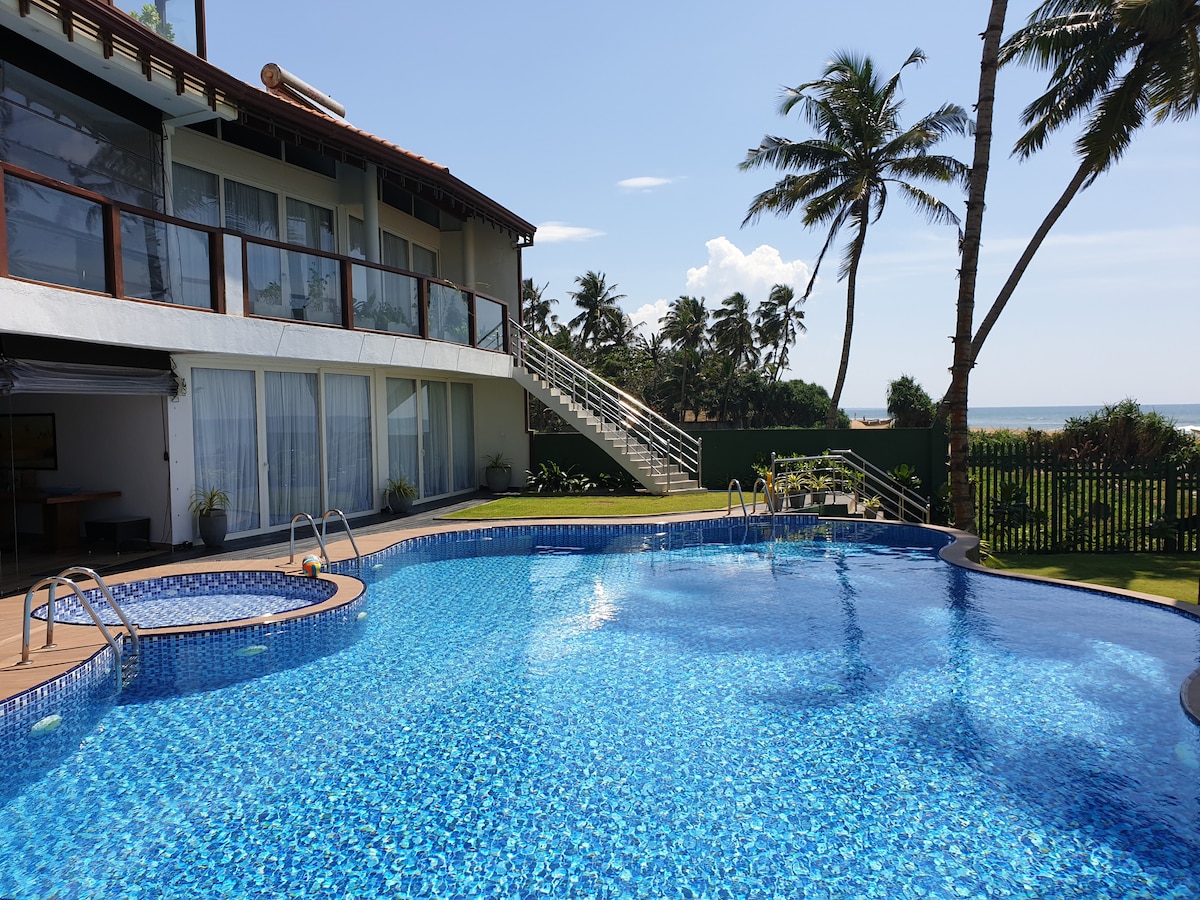 A serene outdoor space features a shimmering swimming pool surrounded by lush greenery. Palm trees provide shade along the edge, and the villa's large glass windows reflect sunny skies, creating a harmonious blend of indoor and outdoor environments.