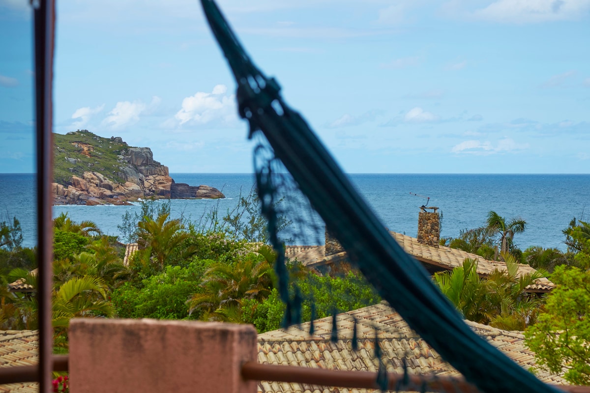 A tranquil view of the ocean is framed by a hammock, with lush greenery and rooftops visible in the foreground. The horizon features distant rocky cliffs, enhancing the serene coastal atmosphere.