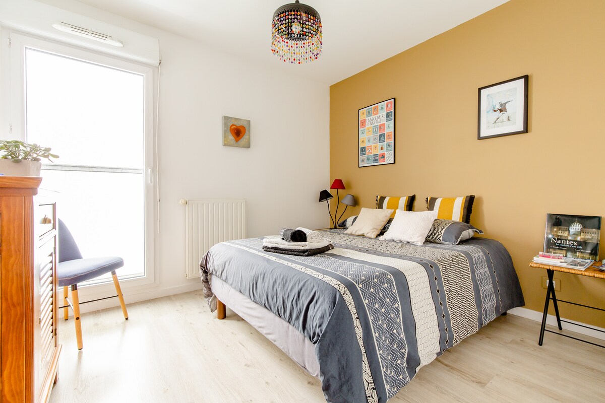 A spacious sleeping area is highlighted by a large bed featuring patterned bedding. A stylish bedside table displays books, while a colorful artwork is framed on the wall. Natural light enters through a large window, complemented by a cozy seating area with a chair.