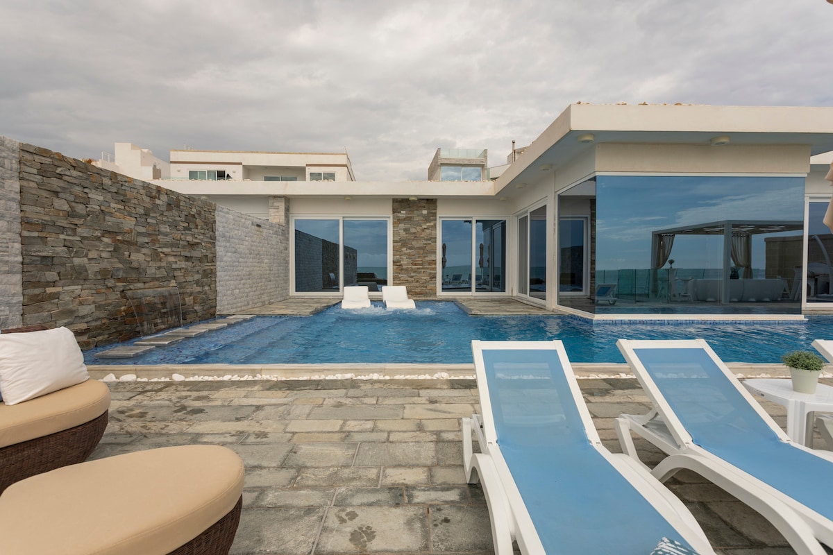 An outdoor pool area features a shimmering blue infinity pool with modern lounge chairs positioned nearby. A stone wall adds texture to the space, while large glass doors of the villa reflect the surrounding serene environment.