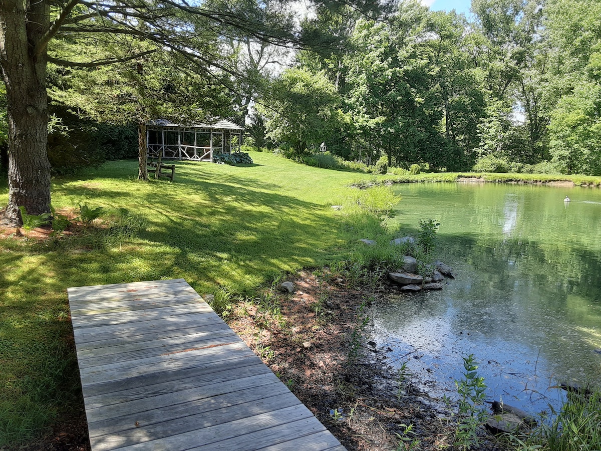 A well-maintained path leads to a serene pond surrounded by lush greenery. A rustic gazebo can be seen in the distance, partially shaded by large trees. The tranquil waters reflect the surrounding nature, enhancing the peaceful ambiance of the property.