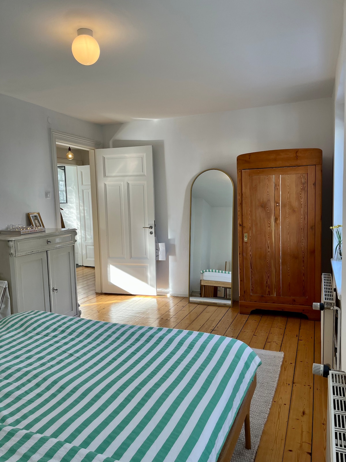 A bright bedroom features a spacious bed with a striped green and white duvet. Natural light filters through the windows, illuminating a wooden wardrobe and a tall mirror. A light fixture hangs from the ceiling, and polished wooden floors add warmth to the room.
