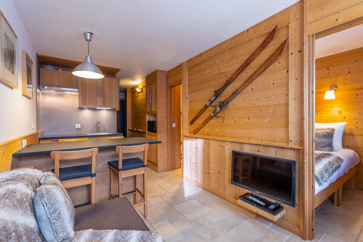 An inviting living area with warm wood paneling is visible, featuring a comfortable sofa and a flat-screen TV. The adjacent kitchen is equipped with modern appliances, and decorative skis are mounted on the wall, adding character. Soft lighting enhances the cozy ambiance.