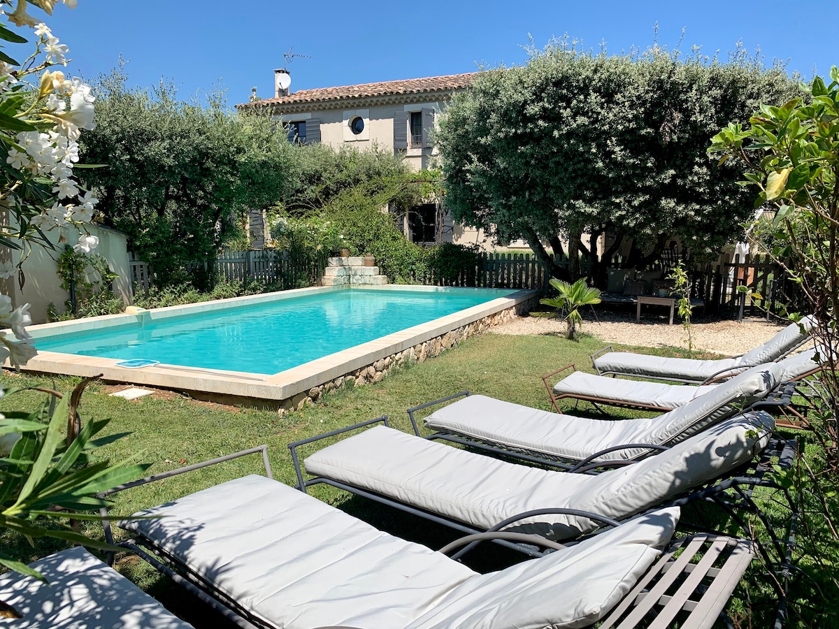 A heated swimming pool is surrounded by a spacious garden featuring several lounging chairs. Lush greenery and flowering plants add a natural touch. The two-story house is visible in the background, set against a clear blue sky.