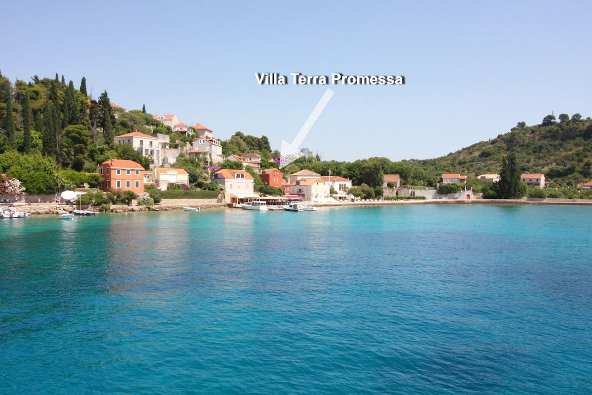 The image captures a picturesque coastal landscape with a view of several Mediterranean-style buildings nestled along the shoreline. The clear blue water reflects the sunlight, enhancing the serene atmosphere of the island. Villa Terra Promessa is highlighted, showcasing its proximity to the sea.